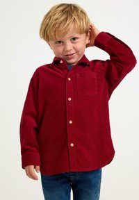 Unge dreng med blondt hår iført en rød skjorte med knapper og blå jeans, stående og rører ved bagsiden af sit hoved.