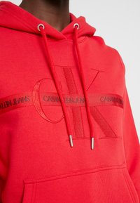 Czerwona bluza z kapturem wykonana z grubej bawełny; na piersi podniesione logo Calvin Klein w kolorach czerwonym i czarnym; srebrne metalowe akcenty na troczkach.