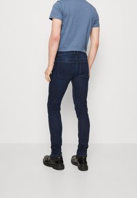 Jeans de mezclilla azul oscuro con un corte ajustado, que cuentan con dos bolsillos traseros y detalles de costura sutiles. Combinados con una camiseta gris de manga corta y zapatos negros.
