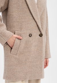 Cappotto in misto lana beige con design doppiopetto, due bottoni e tasche laterali, con una finitura morbida e testurizzata.