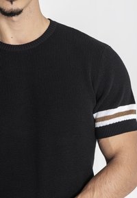 Maglione corto a maniche corte di colore nero, realizzato in maglia con un motivo testurizzato. Presenta un colletto rotondo a coste e tre strisce orizzontali bianche e marroni sulle maniche.