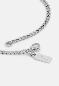 Sølvkjede med runde lenker, en krokfeste og et rektangulært merke med embossed "O & J." Glatt metallisk overflate.