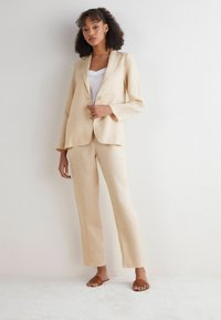 Beige linnen pak met een losse blazer en rechtlopende broek. Gecombineerd met een witte tanktop en bruine slippers. Zachte textuur.