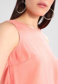 Žena nosící velké zlaté kruhové náušnice a top bez rukávů v korálově růžové barvě, s rovnými tmavě hnědými vlasy viditelnými.