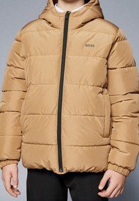 Beige pufferväst med huva, dragkedja och elastiska manschetter. Innehåller horisontellt kviltade sektioner och en logotyp på bröstet.