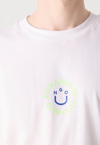 Weißes T-Shirt mit kleinem grünen und blauen grafischen Text "POWERED BY SUNSHINE" und Buchstaben, die in der Nähe der Brust ein Smiley-Gesicht bilden.