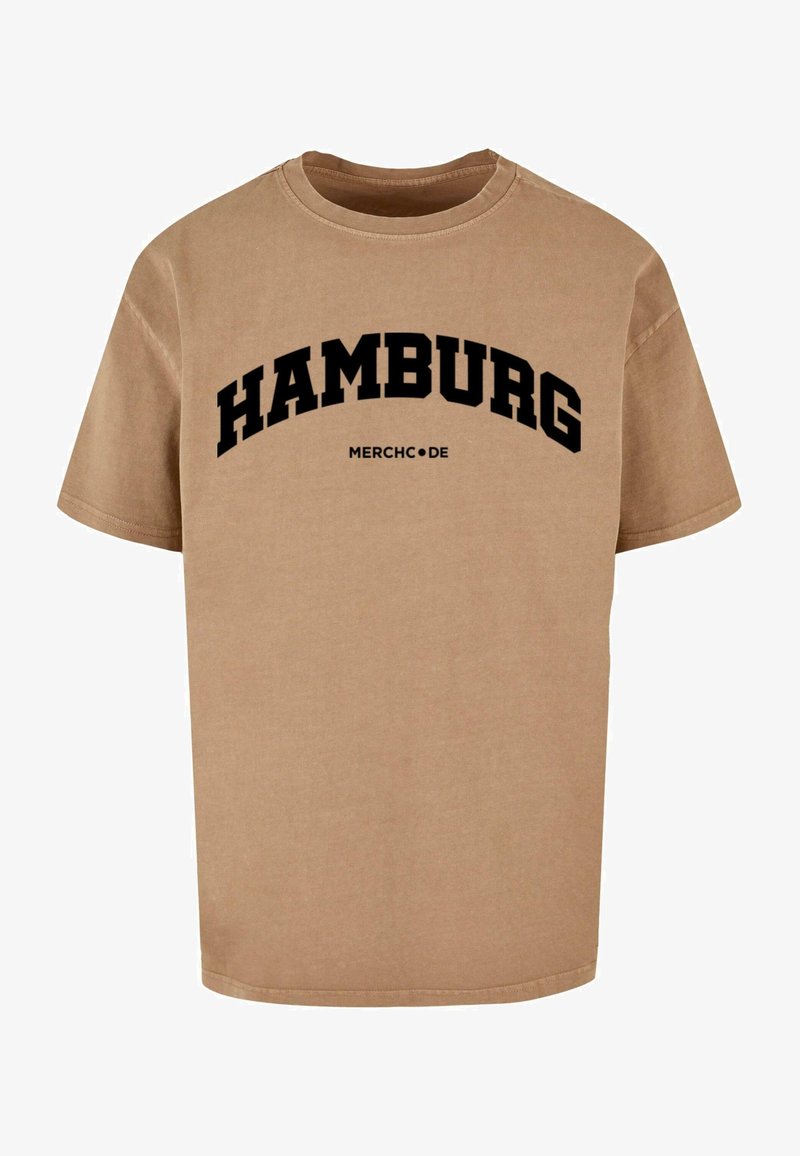 Beige katoenen t-shirt met zwarte tekst die "HAMBURG" in een gebogen ontwerp boven "MERCHC.DE." leest. Korte mouwen en ronde hals.