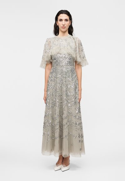 Mujer que lleva un vestido largo beige con lentejuelas florales plateadas y mangas cortas transparentes con volantes, combinado con zapatos blancos puntiagudos.