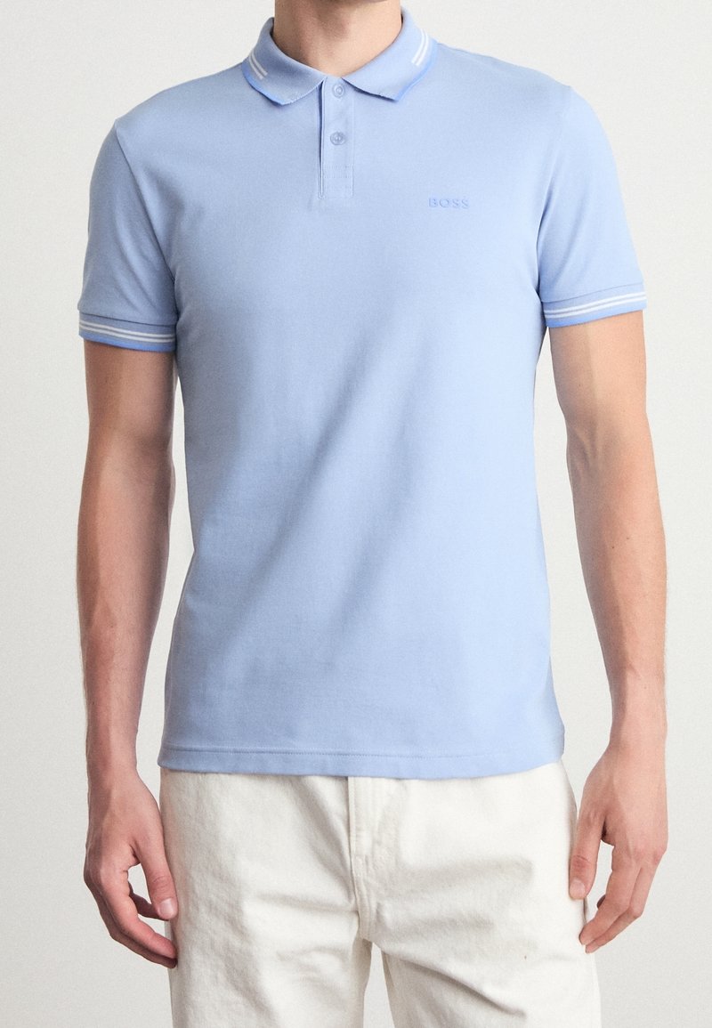 Homme portant un polo bleu clair à manches courtes avec un col et des poignets rayés blancs et un pantalon blanc sur un fond uni.
