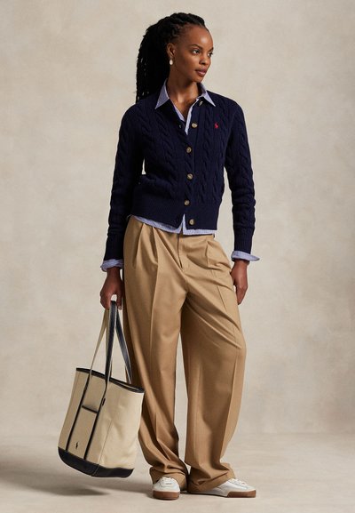 Mujer con el cabello oscuro trenzado que lleva un cárdigan de punto con ochos azul marino sobre una camisa a rayas, pantalones beige de pierna ancha, zapatillas blancas, sosteniendo un bolso tote beige.