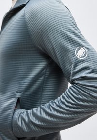 Κοντινό πλάνο ενός ατόμου που φοράει μια υφασμάτινη, γκρι μακριμάνικη ζακέτα με λευκό σήμα μαστόδοντα στο πάνω μέρος του χεριού.