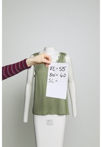 Main tenant une feuille avec des notes manuscrites sur les tailles devant un mannequin sans tête portant un haut vert sans manches sur un fond uni.