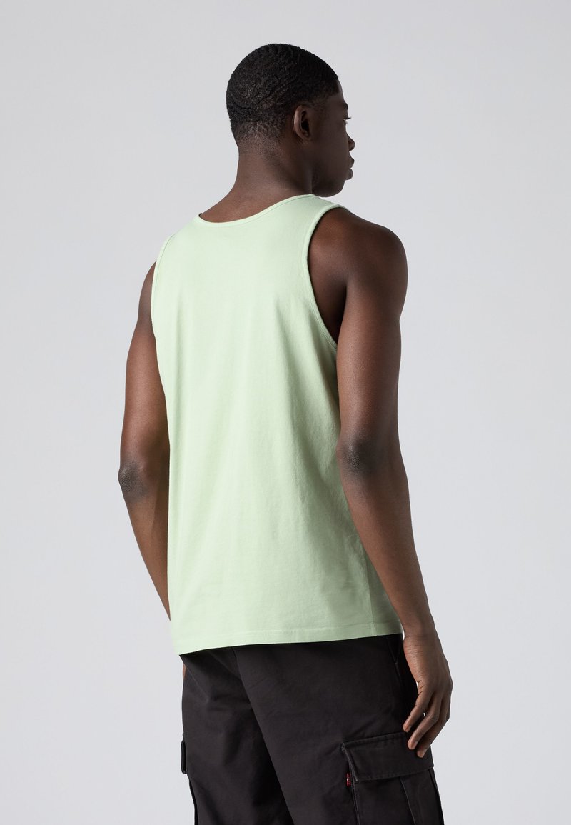 Man draagt een lichtgroen mouwloos hemd en zwarte cargobroek, staand met zijn rug naar de camera tegen een effen grijze achtergrond.