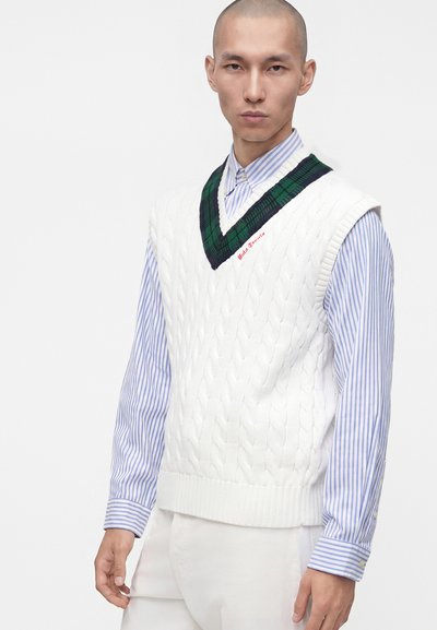 Jeune homme portant un gilet en maille torsadée blanc avec des bordures vertes et noires sur une chemise à rayures bleues et blanches avec boutons.