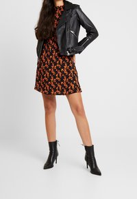 Svart lädermotorcykeljacka över en orange blommönstrad klänning. Modell som bär svarta spetsiga ankelstövlar, står med en hand på höften.