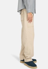 Beige cargo bukser med sidelommer, lige ben-design og synlige syninger, parret med mørkeblå sko med en struktureret sål.