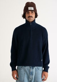 Man wearing a brown knit beanie, navy blue ribbed sweater with a half-zip collar, and light blue denim jeans standing against a plain background.
