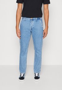 Osoba v světle modrých rovně střihových jeans, černém tričku a černo-modrých teniskách stojí na jednobarevném pozadí.