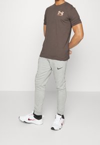 Brun kortärmad T-shirt med logotyp, i kombination med grå avsmalnande träningsbyxor och vita sportskor med svarta och röda detaljer.