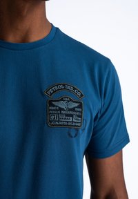 Blauwe katoenen t-shirt met een gestikt label met tekst, embleem en cirkelpatronen. Korte mouwen en een klassieke ronde halslijn.