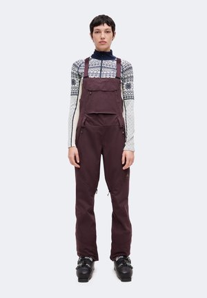 Personne portant un haut à manches longues à motifs, un pantalon de neige à bretelles violet foncé et des bottes de ski noires, debout face à l'avant sur un fond uni.