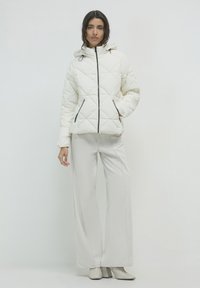 Gepolsterte weiße Daunenjacke mit Kapuze, Reißverschluss vorn und Seitentaschen. Kombiniert mit hellen, gerippten Hosen und beigen Absatzstiefeln.