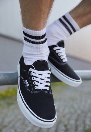 Sneakers neri in tela con lacci bianchi, suola in gomma bianca e cuciture a contrasto. Indossati con calze bianche con strisce nere.