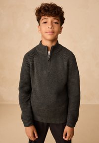 Maglione verde scuro lavorato a maglia con collo alto e cerniera corta, caratterizzato da una texture a coste e polsini, indossato sopra una camicia bianca.