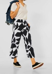 Černobílé vzorované kalhoty s širokými nohavicemi a elastickým pasem, kombinované se světlejším topem s výstřihem do V a černými teniskami, držící džínovou bundu.