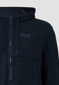 Marine blauwe zip-up hoodie gemaakt van zacht materiaal, met een hoge kraag, voorzakken en een logo patch op de borst. Geborduurde details doorheen.