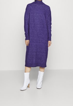 Personne en longue robe en tricot violet et bottes blanches à talons mi-mollet, debout sur un sol gris uni avec un fond clair.
