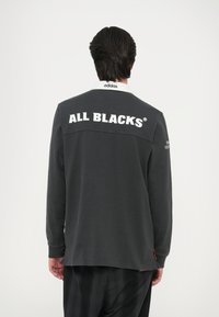 Temno siva majica z dolgimi rokavi z belim ovratnikom; na hrbtni strani nosi veliko napisano "ALL BLACKS" ter subtilno blagovno znamko na rokavu.