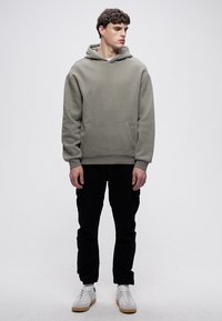 Felpa con cappuccio grigio chiaro con tasca a marsupio, polsini e orlo a coste. Abbinata a pantaloni neri e sneakers bianche, in piedi davanti a uno sfondo semplice.