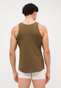 Débardeur sans manches marron avec une texture lisse, ourlet arrondi et grandes emmanchures, porté sur un short blanc, vu de dos.