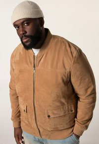 Beige Wildleder Bomberjacke mit einem Frontreißverschluss, zwei unteren Taschen, gerippten Bündchen und einer strukturierten Oberfläche. Kombiniert mit einer leichten Strickmütze.
