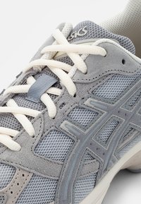 Zapatilla deportiva gris con una mezcla de materiales de malla y ante, cordones en crema y una suela texturizada con un logo distintivo.