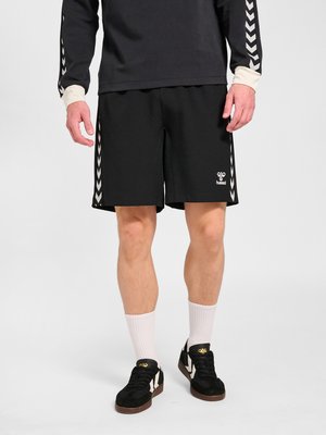 Hombre con pantalones cortos negros con rayas en chevron blancas, zapatillas negras con calcetines blancos, de pie frente a un fondo liso y claro.