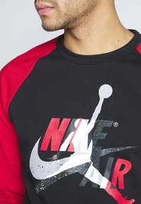 Sudadera negra con mangas raglán rojas. Presenta un gran estampado gráfico de "NIKE AIR" en rojo y blanco con una silueta en gris.