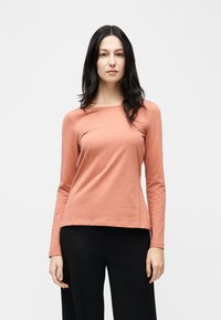 Γυναίκα με μακριά μαύρα μαλλιά που φοράει μπλούζα μακρυμάνικη σε χρώμα σολομού και μαύρο παντελόνι, στέκεται μπροστά σε απλό ανοιχτό φόντο.