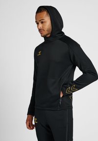 Zwarte sportieve hoodie met een ritszak, voorzien van een gele logo en chevron-ontwerpen op de mouwen. Soepele stof en aansluitend ontwerp.