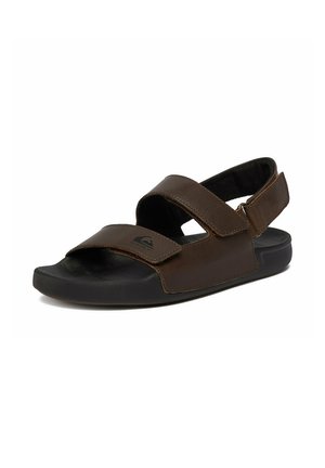 Brun læder sandal med to justerbare remme og en sort polstret sål, vist i en let vinkel på en hvid baggrund.