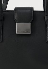 Μαύρη δερμάτινη τσάντα χειρός με ορθογώνιο ασημένιο κούμπωμα που φέρει χαραγμένο το "KARL LAGERFELD" και υφή στην επιφάνειά του.