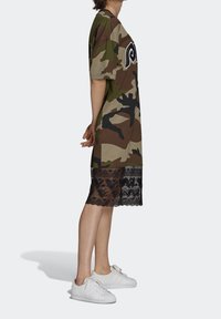 Camouflage print t-shirt dress with short sleeves, featuring a sheer black lace hem. Styled with white sneakers for a casual look.