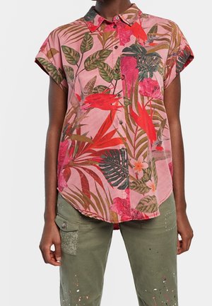 Koszula na krótki rękaw z guzikami, z różowym, czerwonym i zielonym tropikalnym nadrukiem liści i kwiatów, noszona z oliwkowozielonymi spodniami typu cargo z plamami farby.