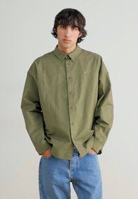 Olivu zaļš pogājams krekls ar apkakli un nelielu logotipu, apvienots ar gaiši zilām džinsu biksēm. Audums izskatās gluds un struktūrēts.