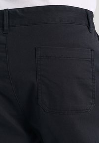 Close-up of black pants showing the back right pocket and waistband with belt loop against a white background.