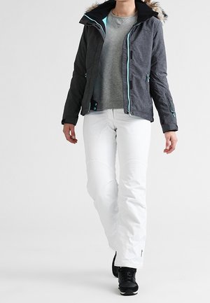 Persona caminando con una chaqueta de invierno oscura con capucha y borde de piel, suéter gris, pantalones blancos y zapatillas negras sobre un fondo liso.