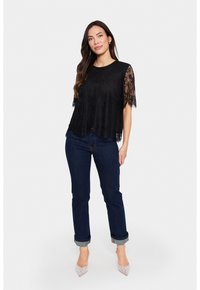 Black top featuring short sheer lace sleeves, soft fabric, and a relaxed fit. Paired with dark blue jeans and pointed-toe shoes.