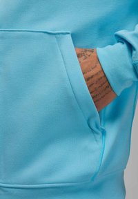 Felpa con cappuccio azzurro chiaro con tasca a marsupio. Presenta un tessuto morbido e dalla trama caratteristica e polsini a coste. Primo piano della tasca e di parte di un braccio tatuato.