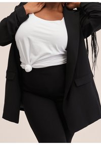 Blazer noir sur un t-shirt blanc noué, associé à un pantalon noir ajusté. Textures lisses, coupe ajustée, sans motifs visibles.